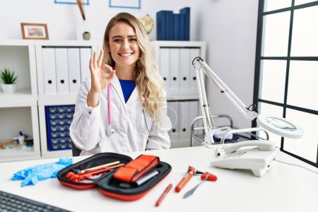 Photo for Young beautiful doctor woman with reflex hammer and medical instruments smiling positive doing ok sign with hand and fingers. successful expression. - Royalty Free Image