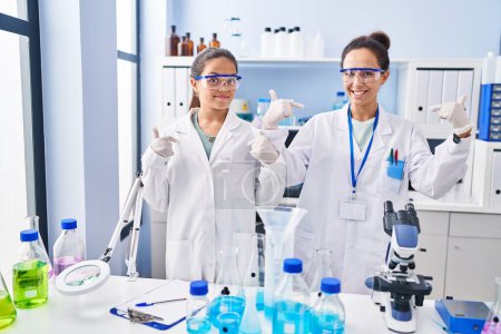 Foto de Young mother and daughter at scientist laboratory looking confident with smile on face, pointing oneself with fingers proud and happy. - Imagen libre de derechos