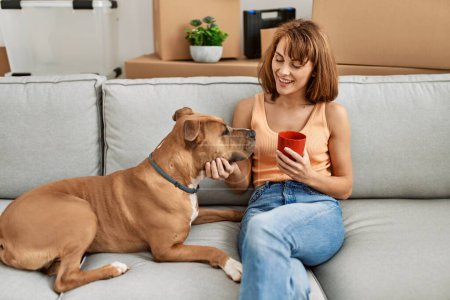 Photo for Young caucasian woman drinking coffee sitting on sofa with dog at home - Royalty Free Image