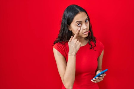 Téléchargez les photos : Young brazilian woman using smartphone over red background pointing to the eye watching you gesture, suspicious expression - en image libre de droit