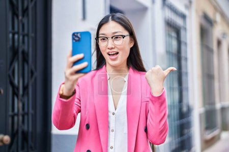 Foto de Mujer joven china haciendo videollamada con smartphone apuntando con el pulgar hacia el lado sonriendo feliz con la boca abierta - Imagen libre de derechos
