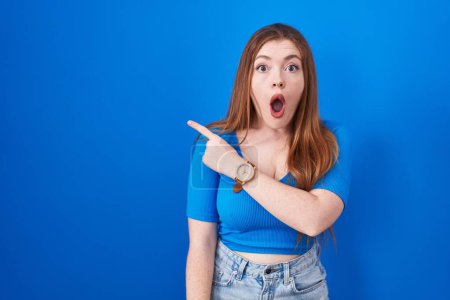 Téléchargez les photos : Redhead woman standing over blue background surprised pointing with finger to the side, open mouth amazed expression. - en image libre de droit