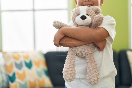 Foto de Adorable niño hispano abrazando oso de peluche de pie en casa - Imagen libre de derechos