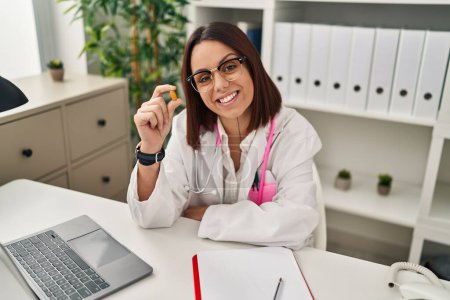 Téléchargez les photos : Young beautiful hispanic woman doctor smiling confident holding pill at clinic - en image libre de droit