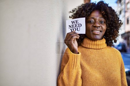 Téléchargez les photos : Afro-américaine femme souriante confiante tenant nous avons besoin de vous papier à la rue - en image libre de droit