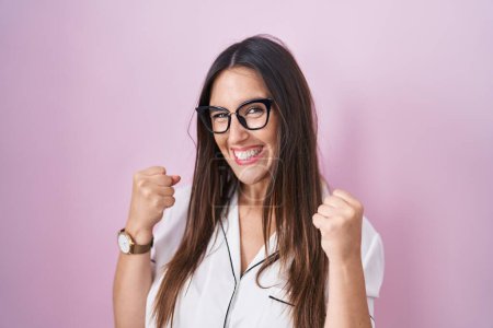 Téléchargez les photos : Jeune femme brune portant des lunettes debout sur fond rose très heureuse et excitée faisant geste gagnant avec les bras levés, souriant et criant pour le succès. concept de célébration. - en image libre de droit
