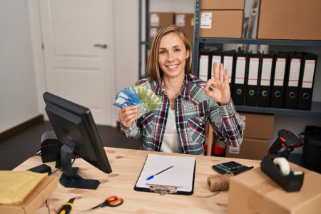 Foto de Young blonde woman working at small business ecommerce holding chilean pesos banknotes doing ok sign with fingers, smiling friendly gesturing excellent symbol - Imagen libre de derechos