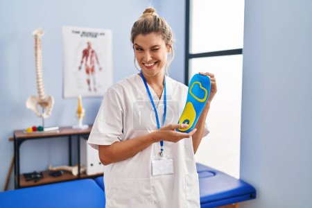 Photo for Young woman holding shoe insole at physiotherapy clinic winking looking at the camera with sexy expression, cheerful and happy face. - Royalty Free Image