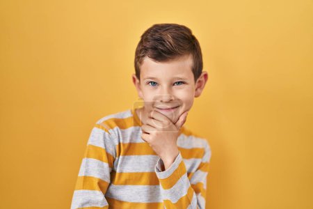 Photo for Young caucasian kid standing over yellow background looking confident at the camera smiling with crossed arms and hand raised on chin. thinking positive. - Royalty Free Image