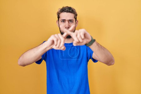 Foto de Hombre hispano con barba de pie sobre fondo amarillo expresión rechazo cruzando dedos haciendo signo negativo - Imagen libre de derechos