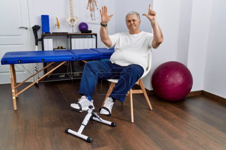 Photo for Senior caucasian man at physiotherapy clinic using pedal exerciser showing and pointing up with fingers number seven while smiling confident and happy. - Royalty Free Image