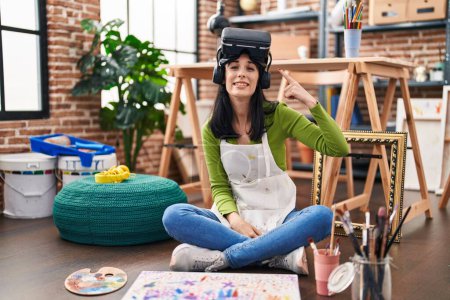 Téléchargez les photos : Hispanic woman painter sitting at art studio wearing virtual reality glasses smiling happy pointing with hand and finger - en image libre de droit
