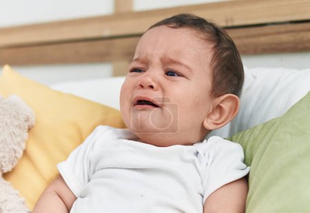 Foto de Adorable bebé hispano sentado en la cama llorando en el dormitorio - Imagen libre de derechos