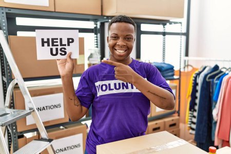 Téléchargez les photos : Jeune homme volontaire africain tenant nous aider bannière souriant heureux pointant avec la main et le doigt - en image libre de droit