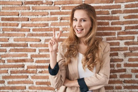 Foto de Beautiful blonde woman standing over bricks wall smiling with happy face winking at the camera doing victory sign. number two. - Imagen libre de derechos