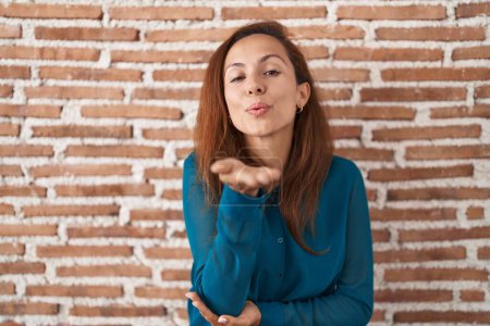 Photo for Brunette woman standing over bricks wall looking at the camera blowing a kiss with hand on air being lovely and sexy. love expression. - Royalty Free Image