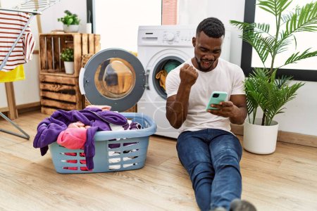 Photo for Young african man doing laundry using smartphone screaming proud, celebrating victory and success very excited with raised arms - Royalty Free Image