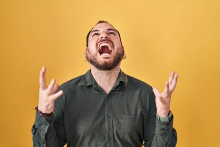 Téléchargez les photos : Plus size hispanic man with beard standing over yellow background crazy and mad shouting and yelling with aggressive expression and arms raised. frustration concept. - en image libre de droit