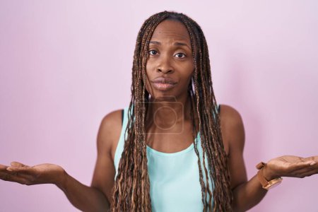 Foto de Mujer afroamericana de pie sobre fondo rosa despistada y confundida con los brazos abiertos, sin concepto de idea. - Imagen libre de derechos