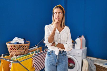 Foto de Mujer rubia joven en la lavandería pensando preocupado por una pregunta, preocupado y nervioso con la mano en la barbilla - Imagen libre de derechos