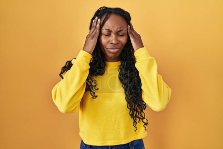Foto de Mujer africana de pie sobre fondo amarillo con la mano en la cabeza para el dolor en la cabeza porque el estrés. padecer migraña. - Imagen libre de derechos