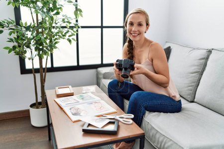 Foto de Joven mujer caucásica haciendo libro de fotos sosteniendo la cámara sentada en el sofá en casa - Imagen libre de derechos
