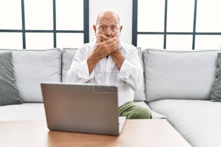 Photo for Senior man using laptop at home sitting on the sofa shocked covering mouth with hands for mistake. secret concept. - Royalty Free Image