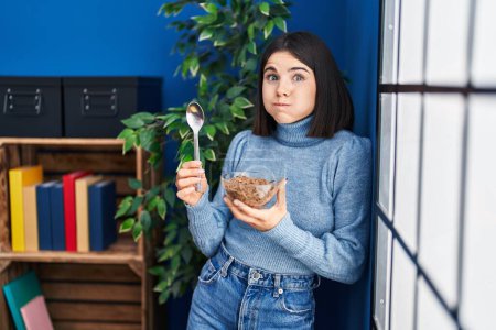Foto de Mujer hispana joven comiendo cereales integrales saludables con cuchara hinchando mejillas con cara divertida. boca inflada con aire, captación de aire. - Imagen libre de derechos