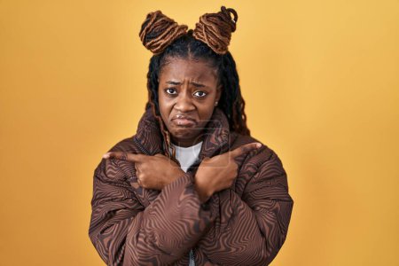 Photo for African woman with braided hair standing over yellow background pointing to both sides with fingers, different direction disagree - Royalty Free Image