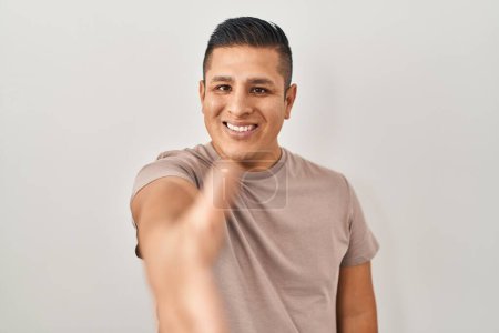 Foto de Joven hispano de pie sobre fondo blanco sonriente amistoso ofreciendo apretón de manos como saludo y bienvenida. negocios exitosos. - Imagen libre de derechos