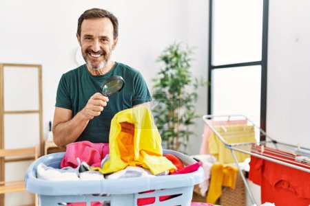Téléchargez les photos : Moyen Age homme hispanique faire la lessive à la recherche de tache sur les vêtements en utilisant loupe à la maison. - en image libre de droit