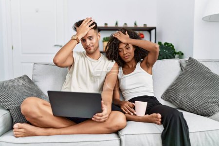 Téléchargez les photos : Jeune couple interracial à l'aide d'un ordinateur portable à la maison assis sur le canapé surpris avec la main sur la tête pour erreur, rappelez-vous erreur. oublié, mauvais concept de mémoire. - en image libre de droit