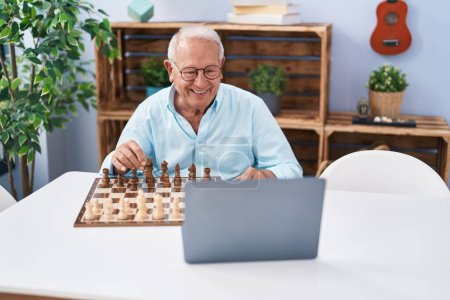 Téléchargez les photos : Senior homme aux cheveux gris souriant confiant jouer jeu d'échecs en ligne à la maison - en image libre de droit