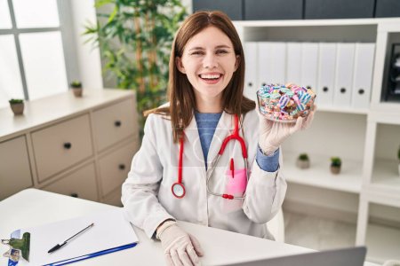 Téléchargez les photos : Jeune femme médecin tenant bonbons bonbons regardant positif et heureux debout et souriant avec un sourire confiant montrant les dents - en image libre de droit