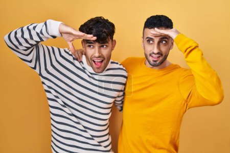 Photo for Young hispanic gay couple standing over yellow background very happy and smiling looking far away with hand over head. searching concept. - Royalty Free Image