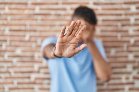 Foto de Brazilian young man standing over brick wall covering eyes with hands and doing stop gesture with sad and fear expression. embarrassed and negative concept. - Imagen libre de derechos