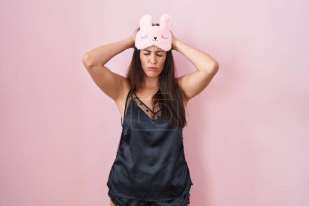 Foto de Young brunette woman wearing sleep mask and pyjama suffering from headache desperate and stressed because pain and migraine. hands on head. - Imagen libre de derechos