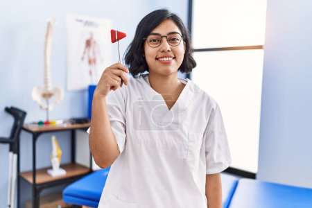 Foto de Young hispanic physiotherapist woman holding reflex hammer looking positive and happy standing and smiling with a confident smile showing teeth - Imagen libre de derechos