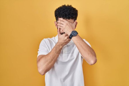 Photo for Arab man standing over yellow background covering eyes and mouth with hands, surprised and shocked. hiding emotion - Royalty Free Image