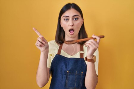 Foto de Hispanic girl eating healthy  wooden spoon surprised pointing with finger to the side, open mouth amazed expression. - Imagen libre de derechos