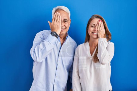 Photo for Middle age hispanic couple standing over blue background covering one eye with hand, confident smile on face and surprise emotion. - Royalty Free Image
