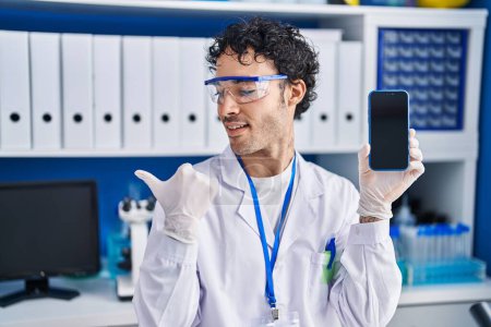 Photo for Hispanic man working at scientist laboratory showing smartphone screen pointing thumb up to the side smiling happy with open mouth - Royalty Free Image