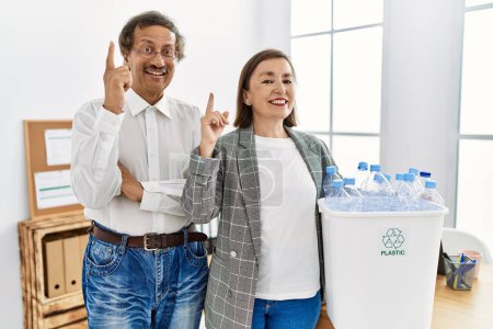 Téléchargez les photos : Couple interracial du Moyen Âge tenant poubelle de recyclage avec des bouteilles en plastique au bureau surpris par une idée ou une question pointant du doigt avec un visage heureux, numéro un - en image libre de droit