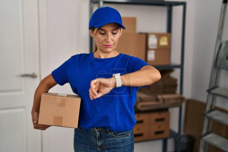 Foto de Middle age brunette woman working wearing delivery uniform and cap checking the time on wrist watch, relaxed and confident - Imagen libre de derechos