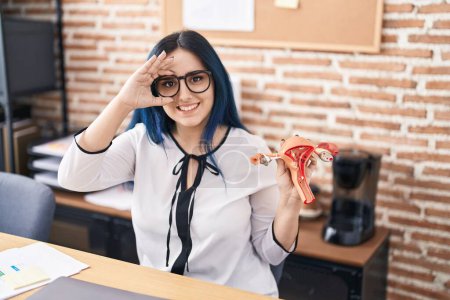 Foto de Chica joven con cabello azul sosteniendo modelo de órgano genital femenino en la oficina quejándose de dolor menstrual sonriendo feliz haciendo signo de ok con la mano en el ojo mirando a través de los dedos - Imagen libre de derechos