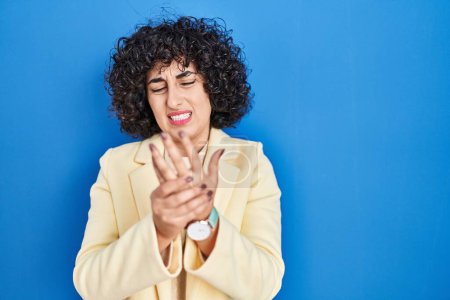 Photo for Young brunette woman with curly hair standing over blue background suffering pain on hands and fingers, arthritis inflammation - Royalty Free Image
