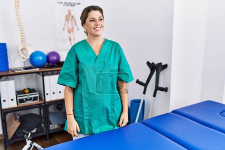 Téléchargez les photos : Young hispanic woman wearing physiotherapist uniform standing at clinic looking away to side with smile on face, natural expression. laughing confident. - en image libre de droit