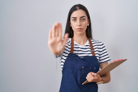 Photo for Young brunette woman wearing professional waitress apron and clipboard with open hand doing stop sign with serious and confident expression, defense gesture - Royalty Free Image