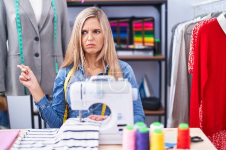 Photo for Blonde woman dressmaker designer using sew machine pointing aside worried and nervous with forefinger, concerned and surprised expression - Royalty Free Image