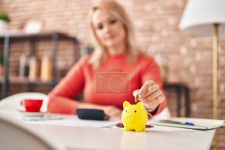 Photo for Young blonde woman inserting coin on piggy bank at home - Royalty Free Image